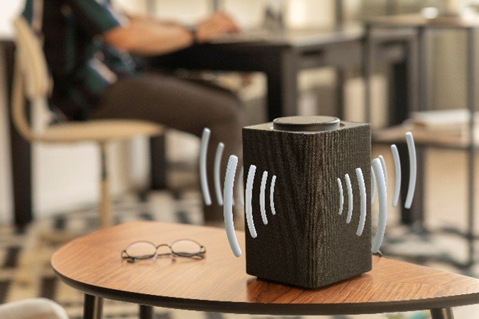 A dark wood-grain wireless speaker sits on a coffee table with a pair of eyeglasses nearby.